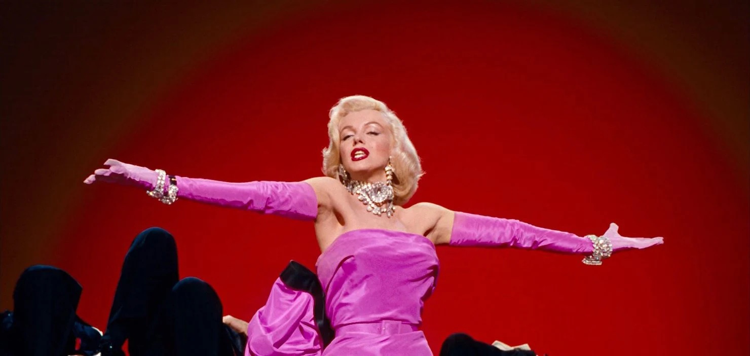 Photo of Marilyn Monroe performing with her arms extended while wearing a vibrant pink strapless outfit and long pink gloves.