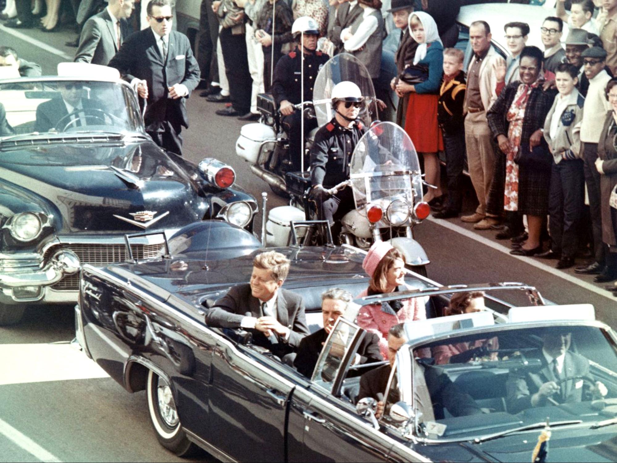 Photo of John and Jackie Kennedy in a black convertible with the top down processing in a motorcade on the day of his assassination.