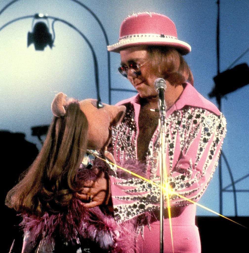 Elton John in a glittery pink jacket and hat embracing Miss Piggy on a stage.