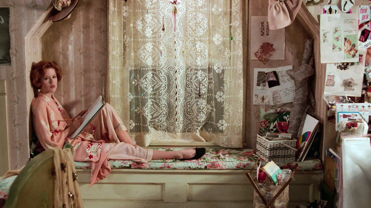 A still from the movie “Pretty in Pink,” featuring a woman with red hair sitting in a nook of her bedroom window staring off, while wearing a pink outfit.