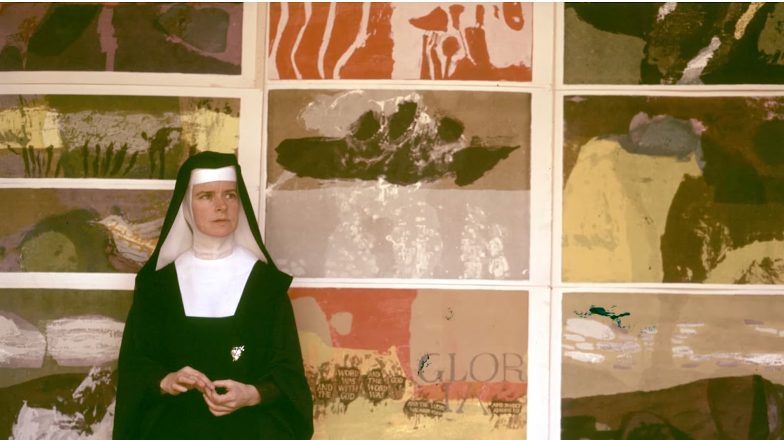 Corita Kent dressed in a habit, standing in front of a wall featuring her artwork