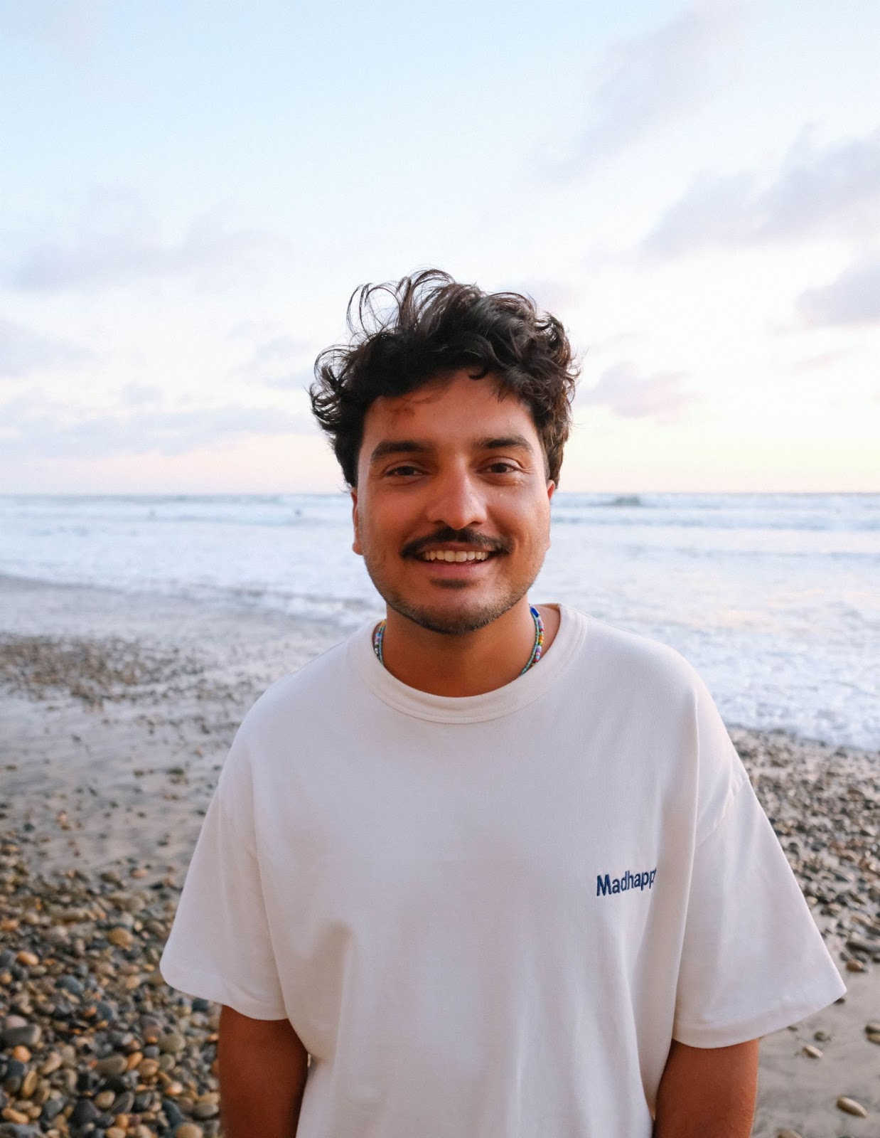 Deepak Kanda, smiling in a white t-shirt on a beach at sunset