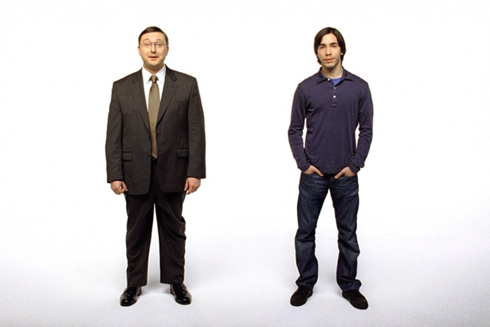Photo of actors John Hodgman and Justin Long standing in a white-wall studio, both addressing the camera. John wears a brown suit, tie, and glasses, while Justin wears jeans and an untucked polo, his hands in his pockets in a casual stance.