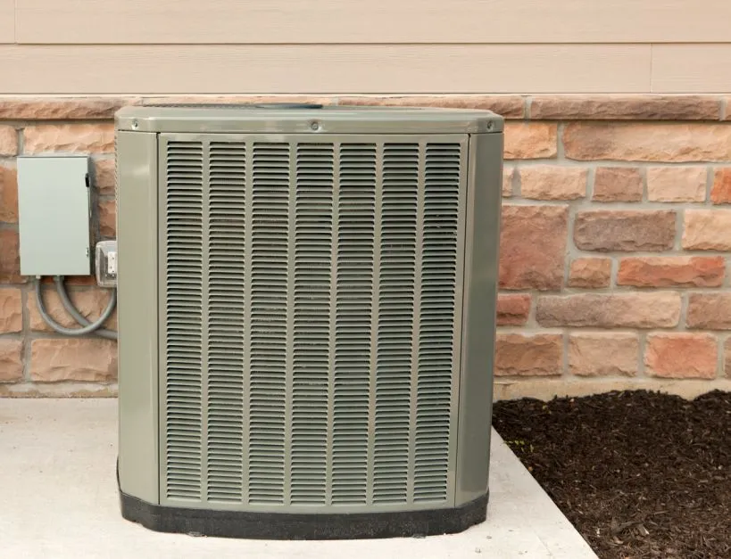 A gray heat pump unit sits on a concrete pad next to a house with a stone and tan siding exterior.