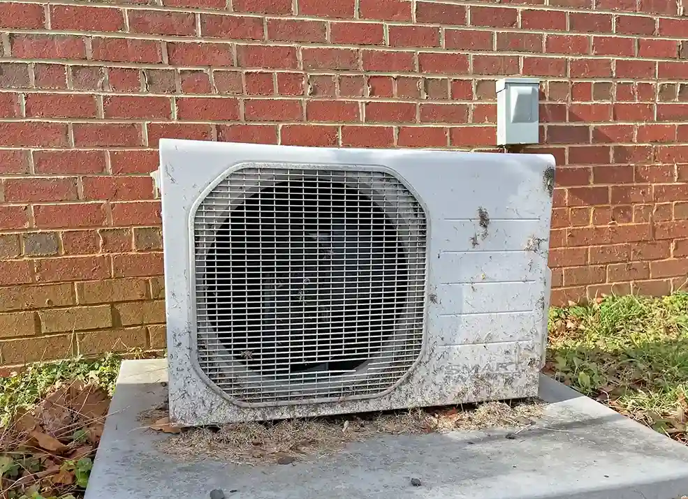 An old and dirty mini-split outdoor unit sitting on a concrete pad in front of a brick wall.