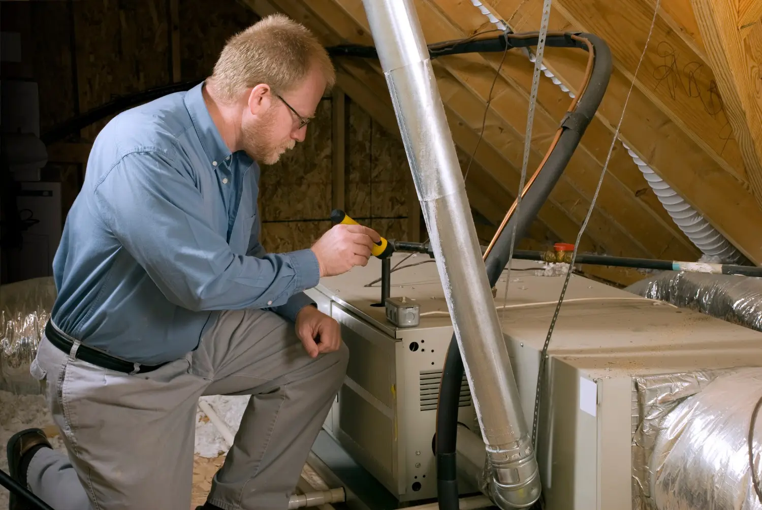 A male technician wearing glasses and a blue shirt is kneeling in a dusty attic, using a yellow tool or light to inspect an HVAC air handler or furnace unit. Large metal ductwork and a black insulated pipe run around the unit, with the wooden roof trusses visible in the background.