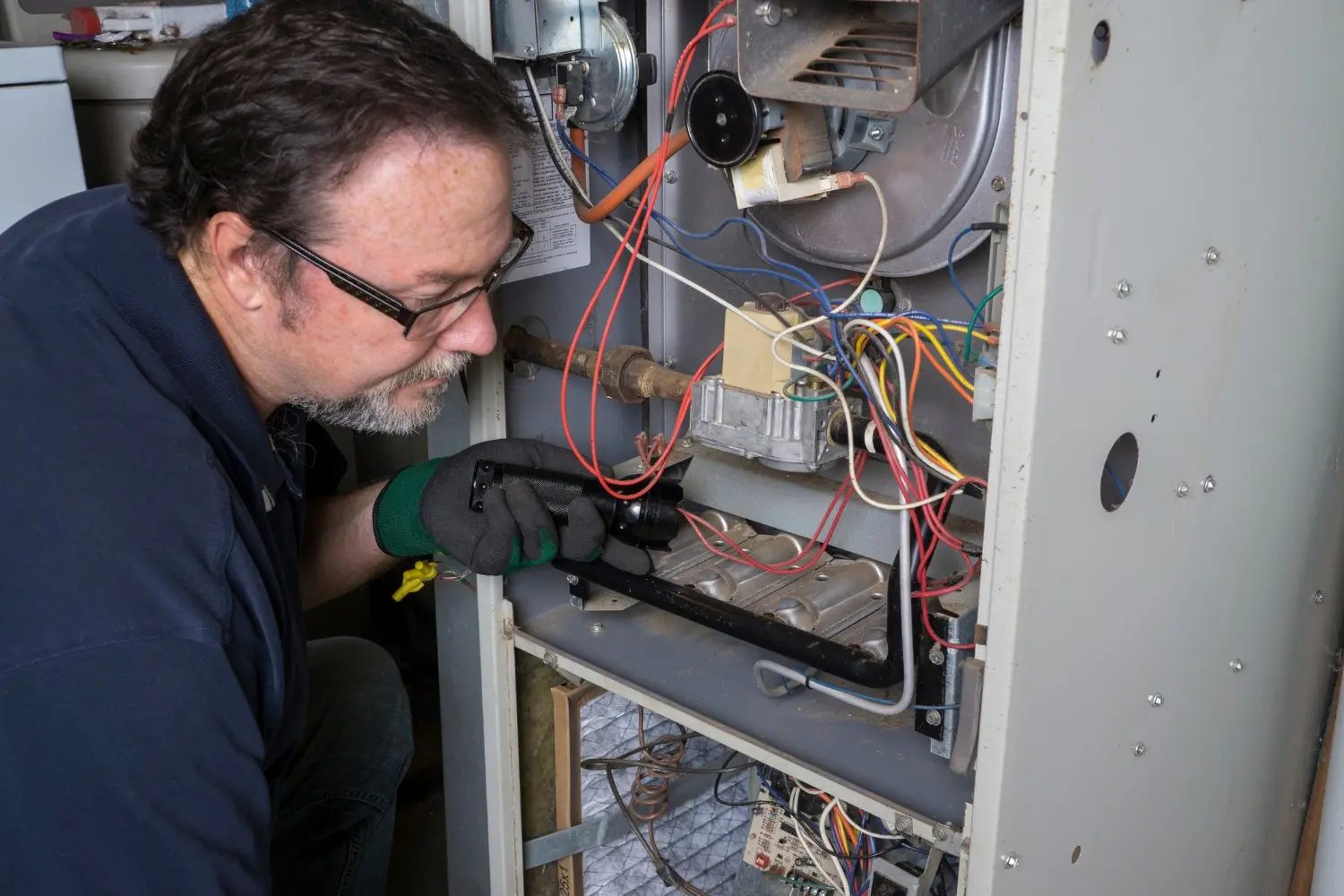 A male HVAC technician with a beard and glasses inspects the burner assembly and internal components of an old light-gray furnace, using a flashlight in his gloved hand.