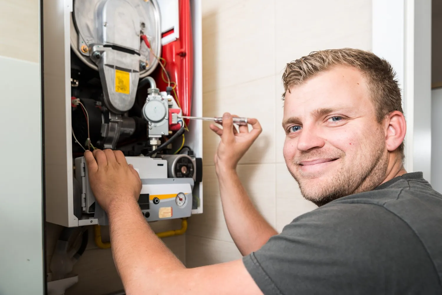 A smiling male technician with a light beard is shown from the side, actively working on the internal components of an open, wall-mounted boiler. He is holding a screwdriver and adjusting a part. The heater features a red section and a control panel, all against a neutral tiled wall.