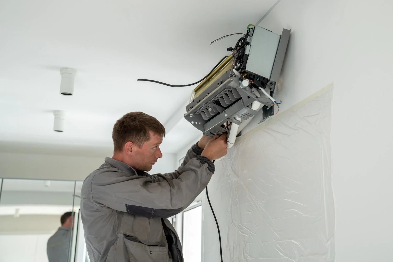 a male technician in a gray uniform working on the installation of a mini-split AC indoor unit on a white wall. The unit's cover is fully removed, exposing the internal components, and the technician is focused on connecting or adjusting the drain line and wiring that are emerging from the wall. A protective plastic sheet is taped to the wall directly beneath the unit to prevent damage or staining during the work. The bright room has white walls and ceiling lights, and a mirror is visible on the left side, reflecting the technician.