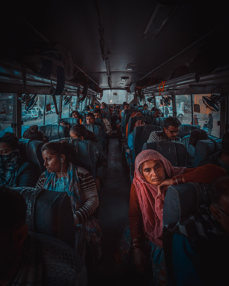Inside view of a crowded bus with Indian passengers seated, some looking out the window and a woman in a pink headscarf resting her head on her hand.