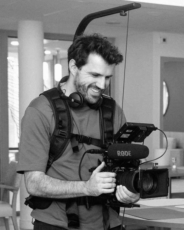 Rémy Pujol operating a professional video camera rig indoors, wearing headphones around his neck.