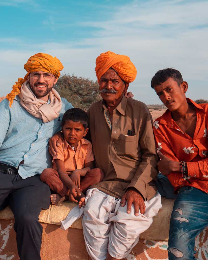 Rémy seated outside among locals — two in orange turbans, one child in a peach shirt, and a man in a red floral shirt.