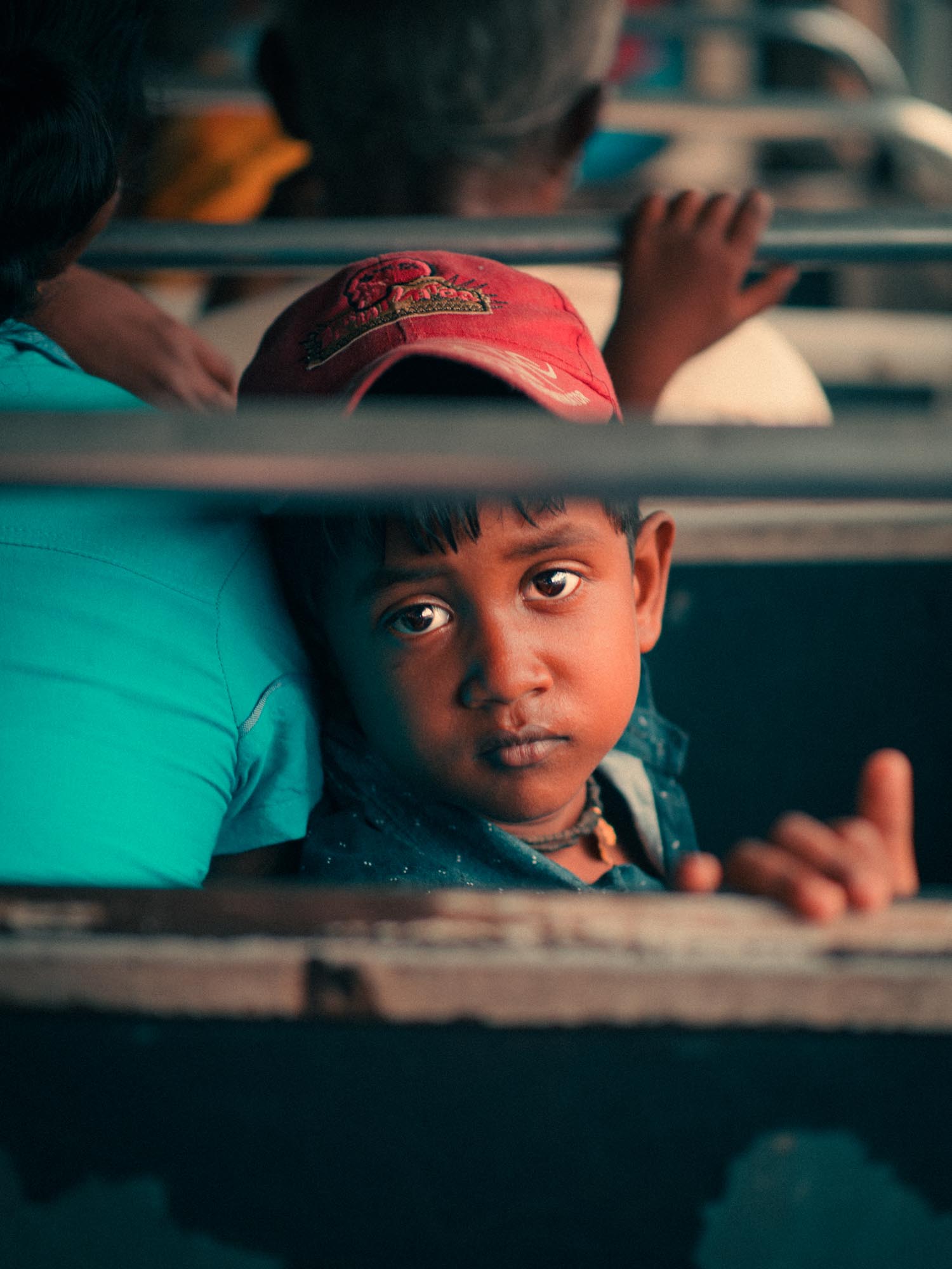 Young boy wearing a red cap looking solemnly through metal bars, leaning on a person in a teal shirt.