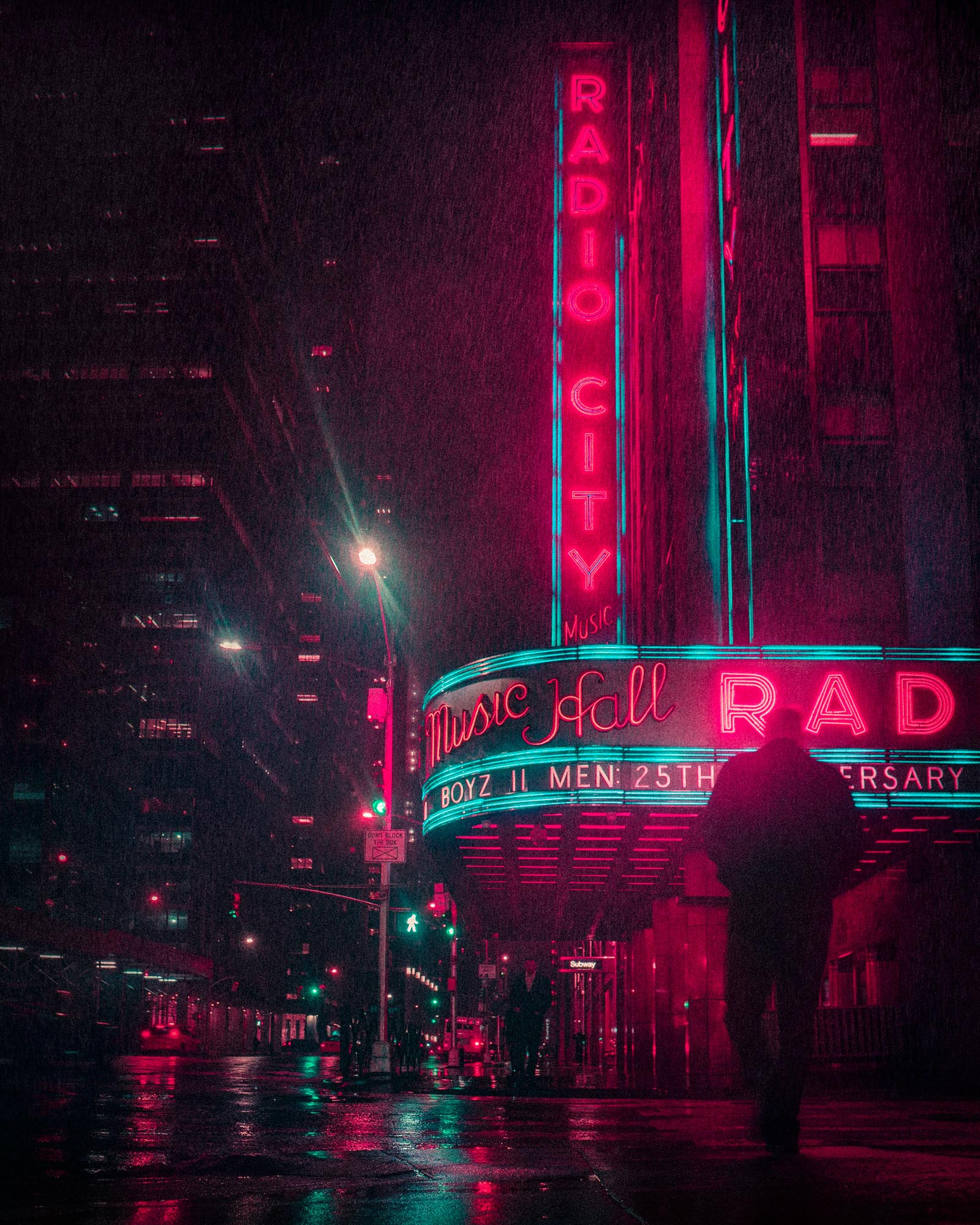 Nighttime street view of Radio City Music Hall illuminated by neon pink and blue lights with a silhouetted person walking in the foreground.