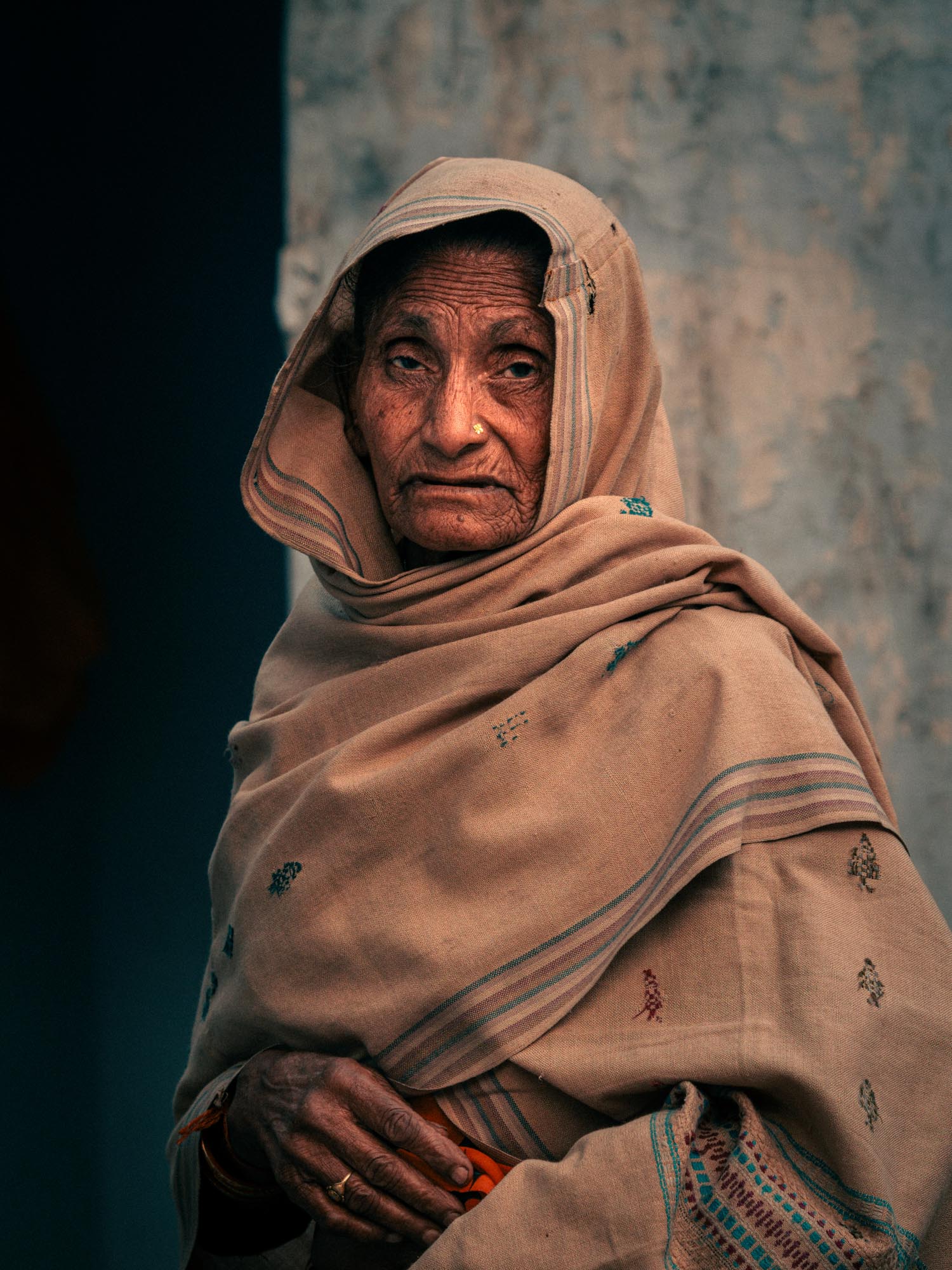 Portrait of an elderly woman wearing a beige shawl with traditional embroidery and a nose ring, standing against a blurred wall background.