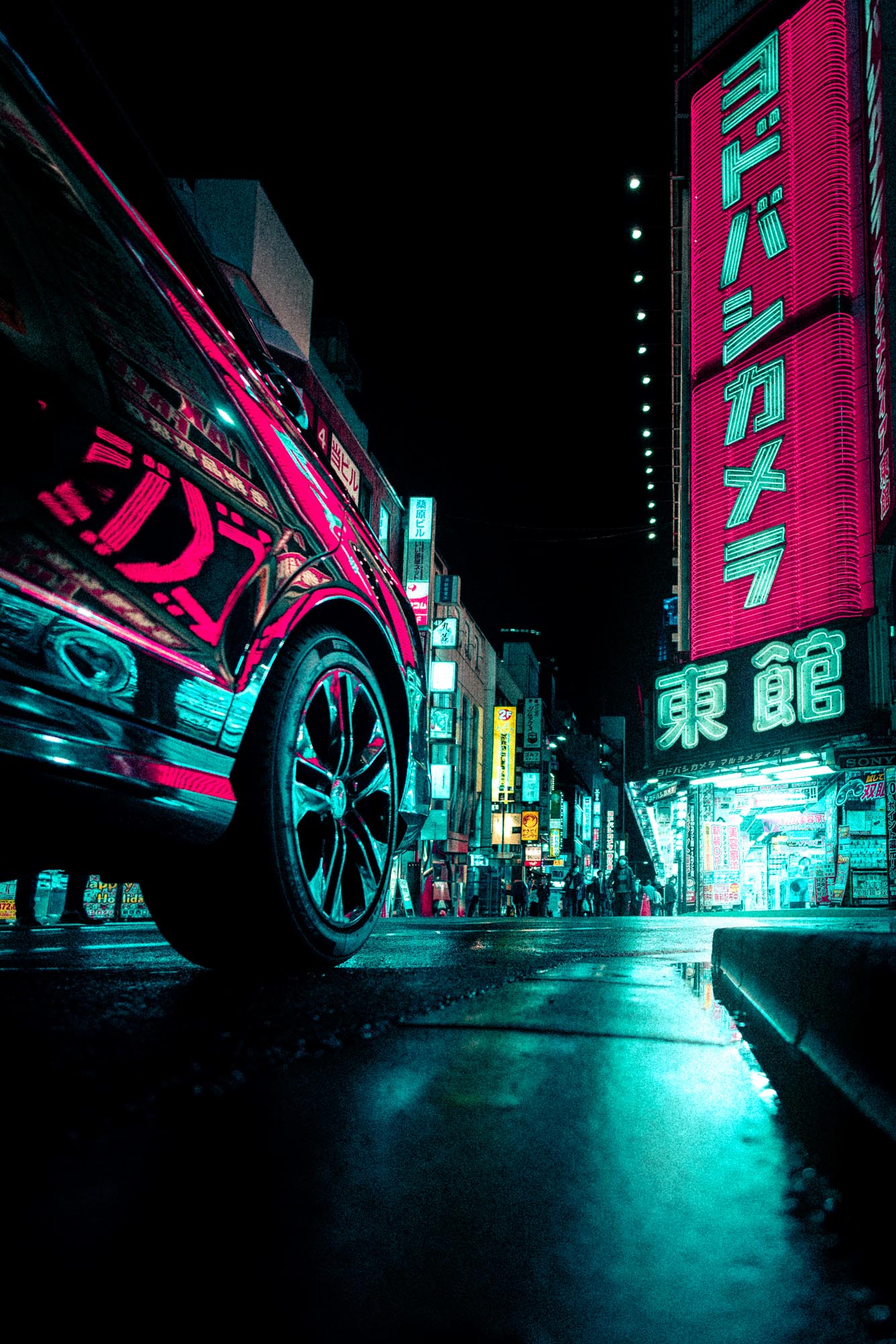 Low-angle view of a street at night with a close-up of a car wheel and neon-lit signs in Japanese on buildings.
