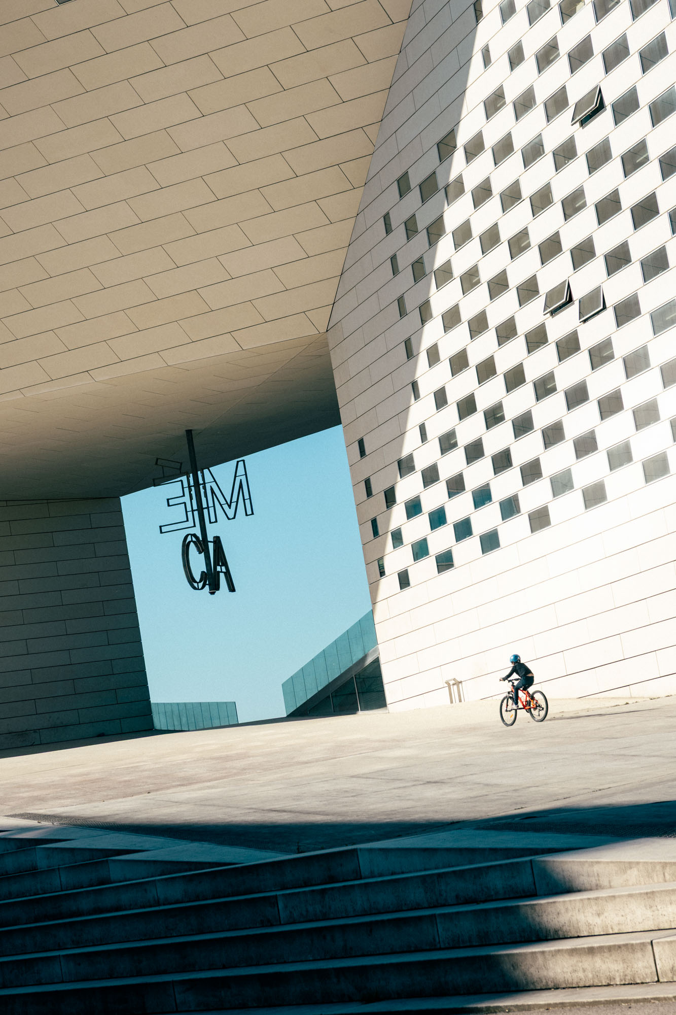 Person wearing a helmet riding a bicycle near a modern building with patterned windows and an outdoor neon sign reading 'ME CA'.