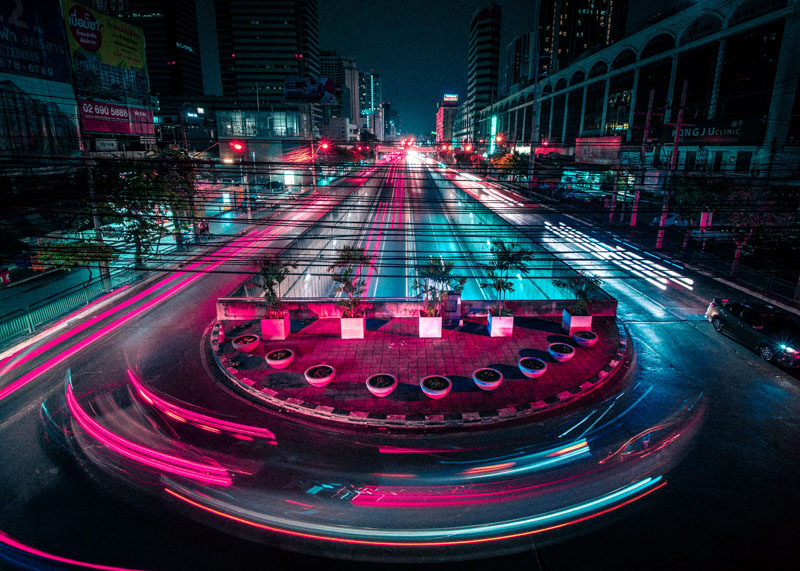 Nighttime city street with a roundabout center featuring plants, illuminated by pink and blue light trails from moving vehicles.