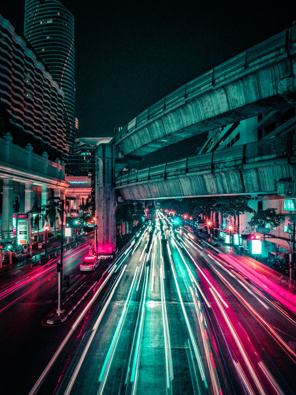 Night cityscape with long-exposure light trails from vehicles on a multi-lane street under elevated concrete train tracks.