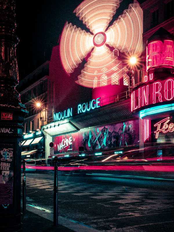 Night scene of Moulin Rouge with illuminated spinning windmill and neon signs on a city street.