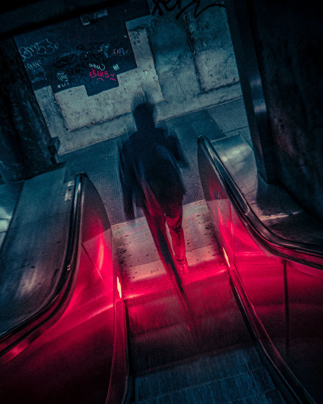 Blurred figure descending an escalator illuminated by red lights in a dim, graffiti-covered urban environment.
