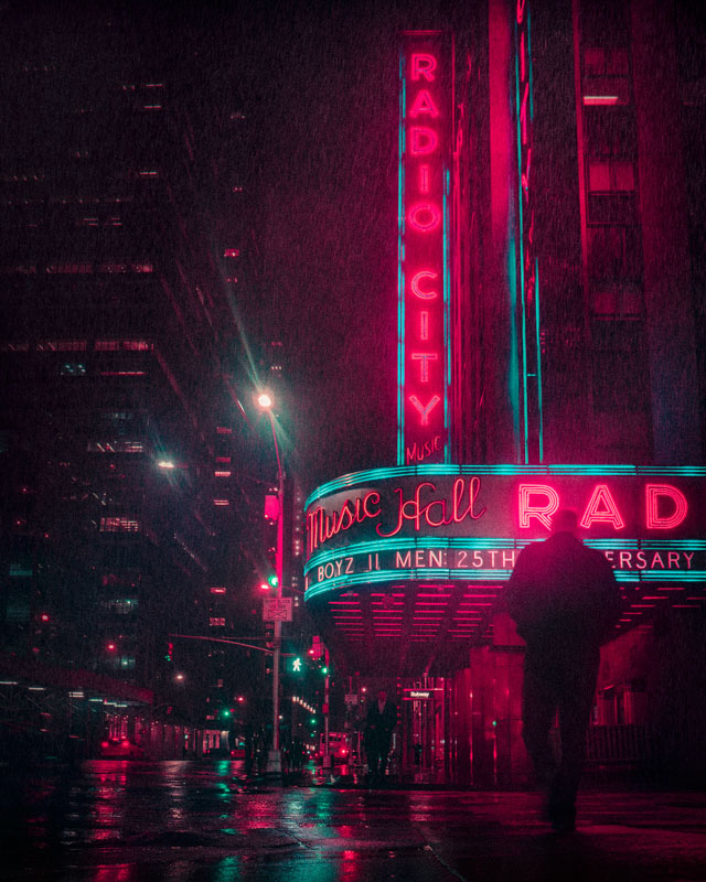 Rainy night street view of Radio City Music Hall with neon pink and blue lights and people walking.