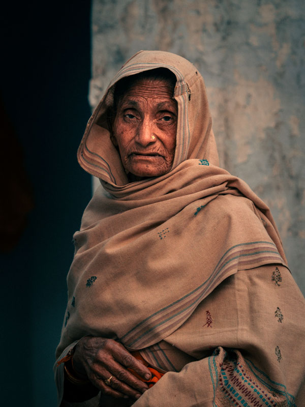 Elderly woman wearing a beige shawl with small embroidered patterns, standing against a textured wall.