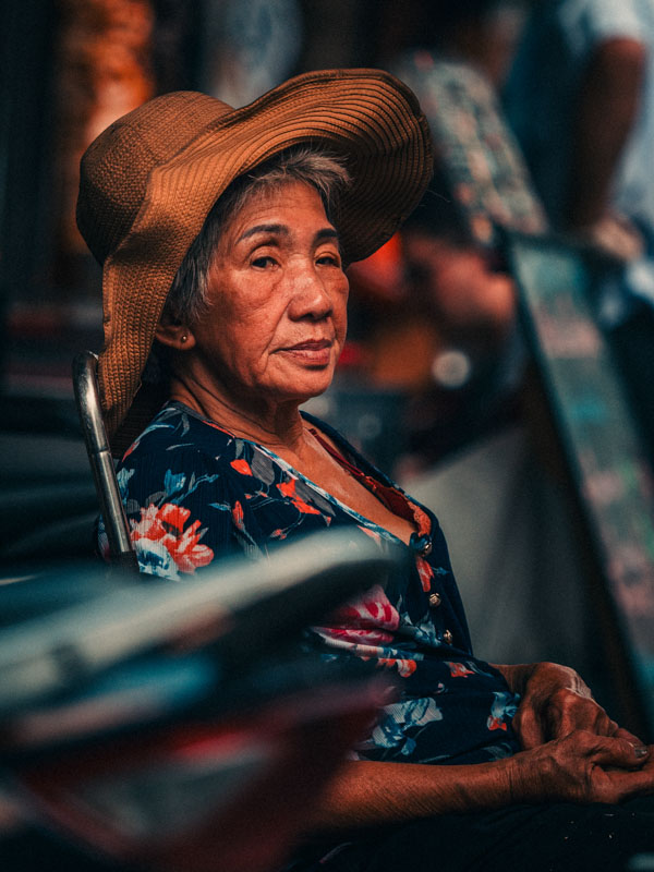 Elderly woman wearing a large brown sunhat and floral blouse sitting in a chair with a contemplative expression.