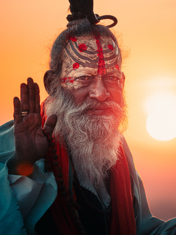 Elderly man with painted face, long white beard, and traditional attire raising his hand in blessing against a sunset background.