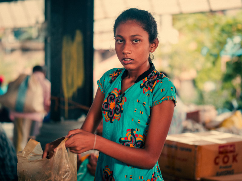 Young girl in a teal dress with colorful patterns holding a plastic bag in a busy market setting.
