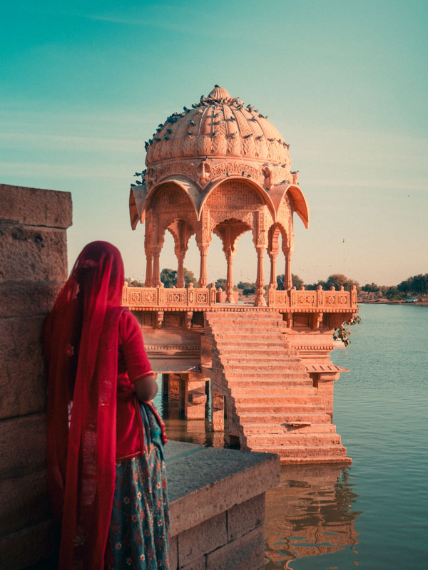 Woman in traditional Indian attire looking at an ornately carved sandstone pavilion on steps leading into a calm body of water.