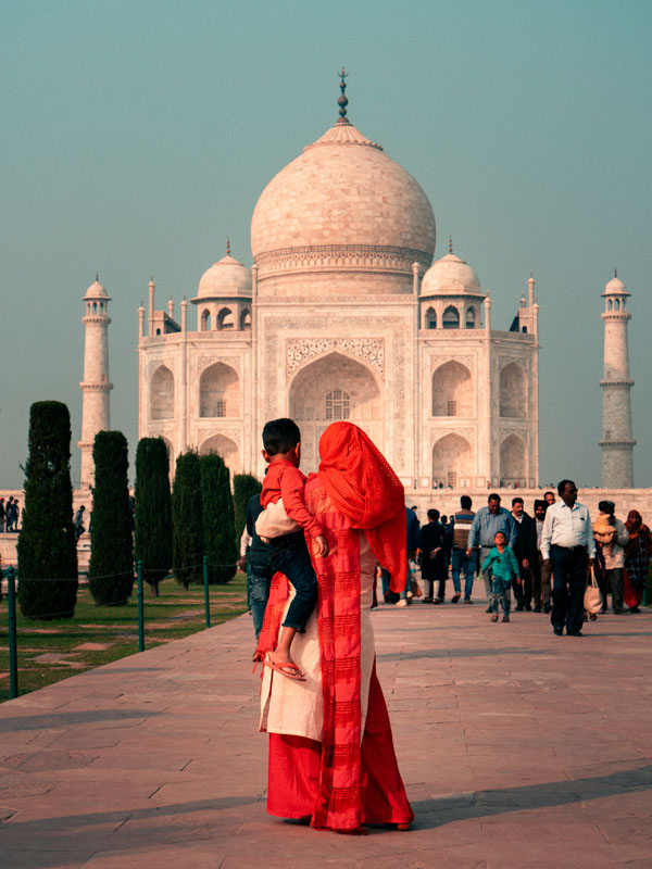 A woman in a red sari carrying a child, standing on a pathway facing the Taj Mahal with other visitors around.