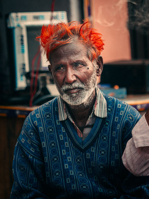 Elderly man with bright orange hair and a patterned blue sweater sitting indoors.