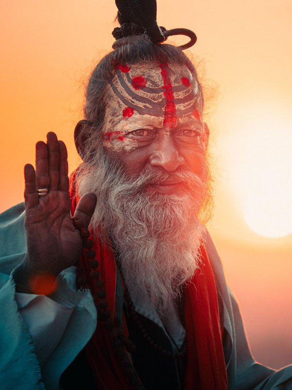 Elderly man with painted face and long white beard raising his hand in blessing at sunset.