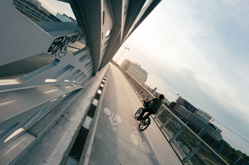 Person riding a bicycle on an elevated bike path with city buildings and cranes in the background at sunset.