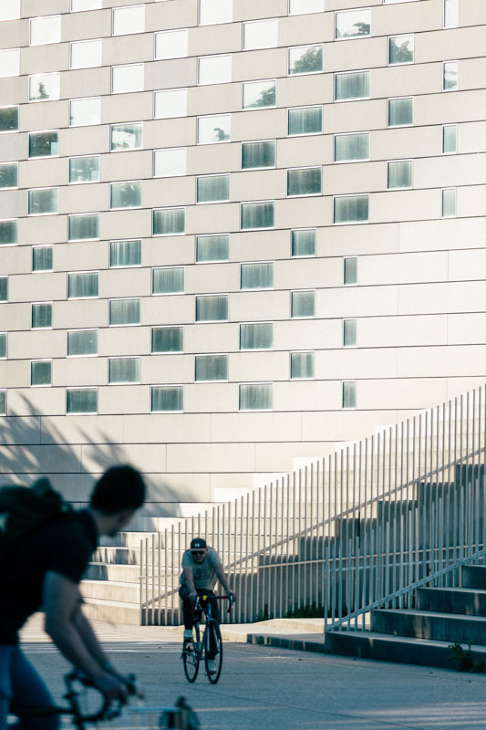 Two cyclists riding near a modern building with a patterned facade and metal railings along stairs.