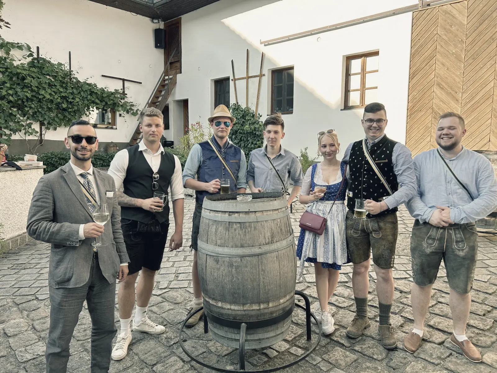 Eine Gruppe von sieben Personen steht um ein großes Holzfass auf einem gepflasterten Innenhof. Die meisten tragen traditionelle Trachten, darunter Lederhosen und ein Dirndl, einige halten Bier- oder Weingläser in der Hand. Im Hintergrund sind ein weißes Haus mit Fenstern und eine Holztreppe zu sehen.