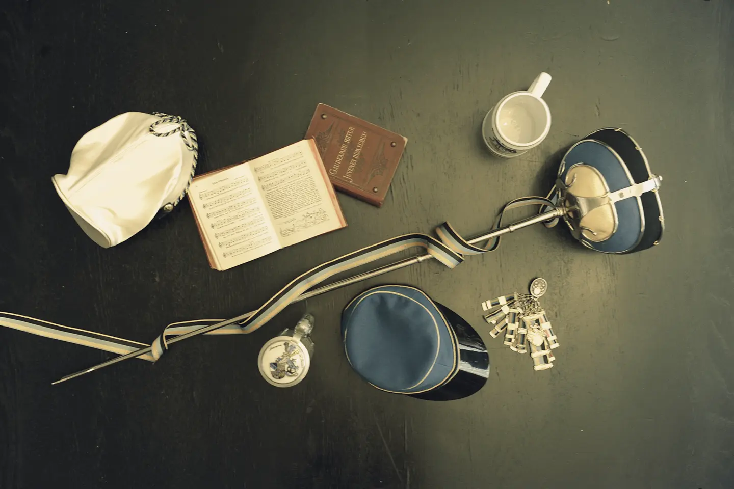 Auf einem dunklen Tisch liegen verschiedene historische Objekte: eine weiße Kapuze, ein geöffnetes Buch mit Noten und Text, eine kleine silberne Flasche, zwei blaue Mützen mit goldener Umrandung, ein silberner Trinkbecher mit Henkel und ein langer silberner Stab mit einer weißen, blau umrandeten Schutzhülle und einer bunten Kette mit Anhängern.