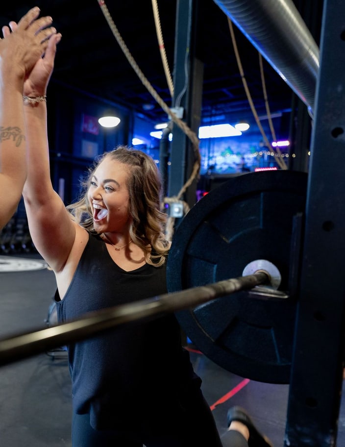 Two BLKOPS FITNESS members smiling and raising their arms in a high five next to a barbell in the gym