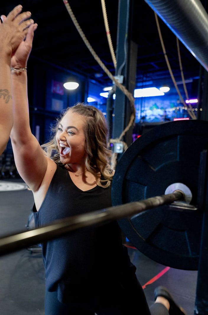 Two BLKOPS FITNESS members smiling and raising their arms in a high five next to a barbell in the gym