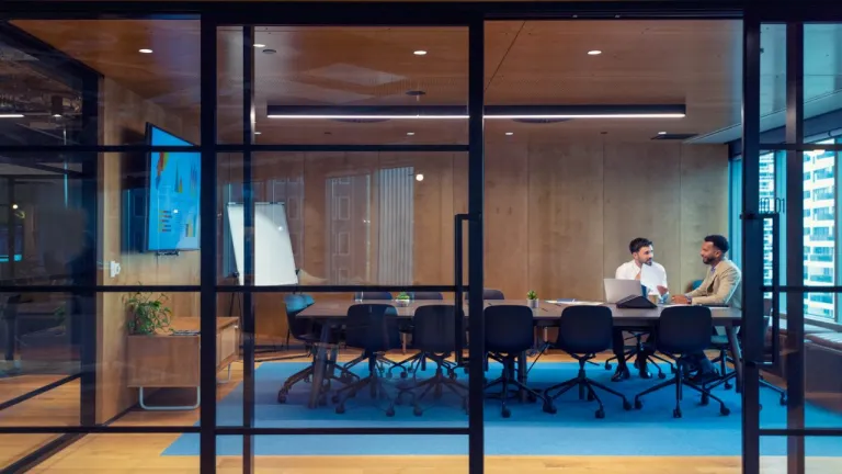 Two men seated at a conference table in a modern office meeting room with glass walls and a blue carpet.