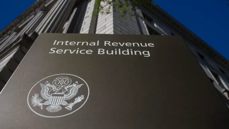 Sign reading 'Internal Revenue Service Building' with the IRS emblem, in front of a tall building under a clear blue sky.
