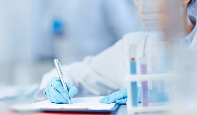 Lab technician wearing blue gloves writing notes with a pen beside test tubes.