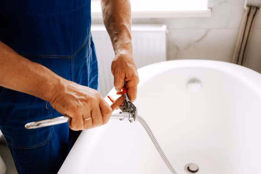 A skilled plumber kneels in front of a kitchen sink. Dressed in a navy blue polo with the logo "Raymond's Plumbing," 