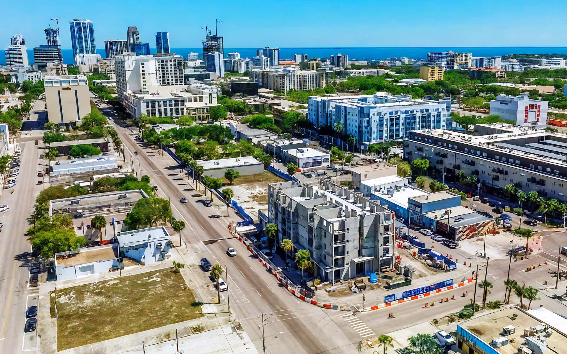 A vibrant urban landscape unfolds in this aerial view, showcasing a bustling area dotted with modern high-rise buildings that reach towards a pristine blue sky. The foreground features an active construction site, with a partially completed structure at its center, surrounded by construction barriers