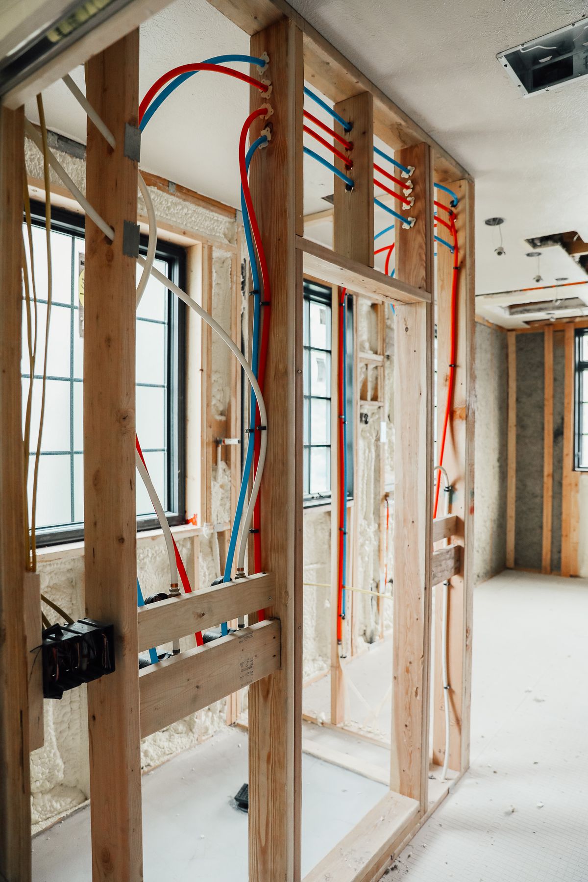 Interior of a building under construction showing exposed wooden framing with blue and red plumbing pipes and partially insulated walls.