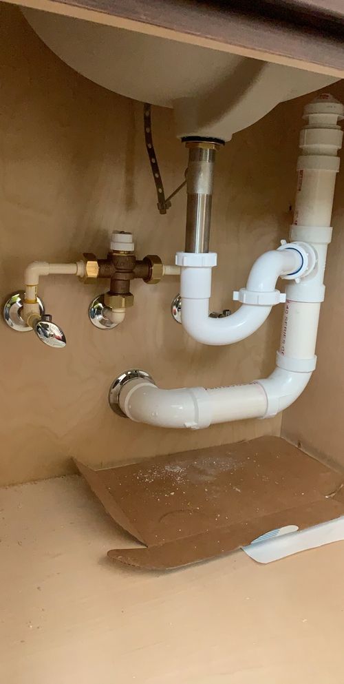 Plumbing pipes and valves under a sink inside a wooden cabinet with a piece of cardboard on the cabinet floor.
