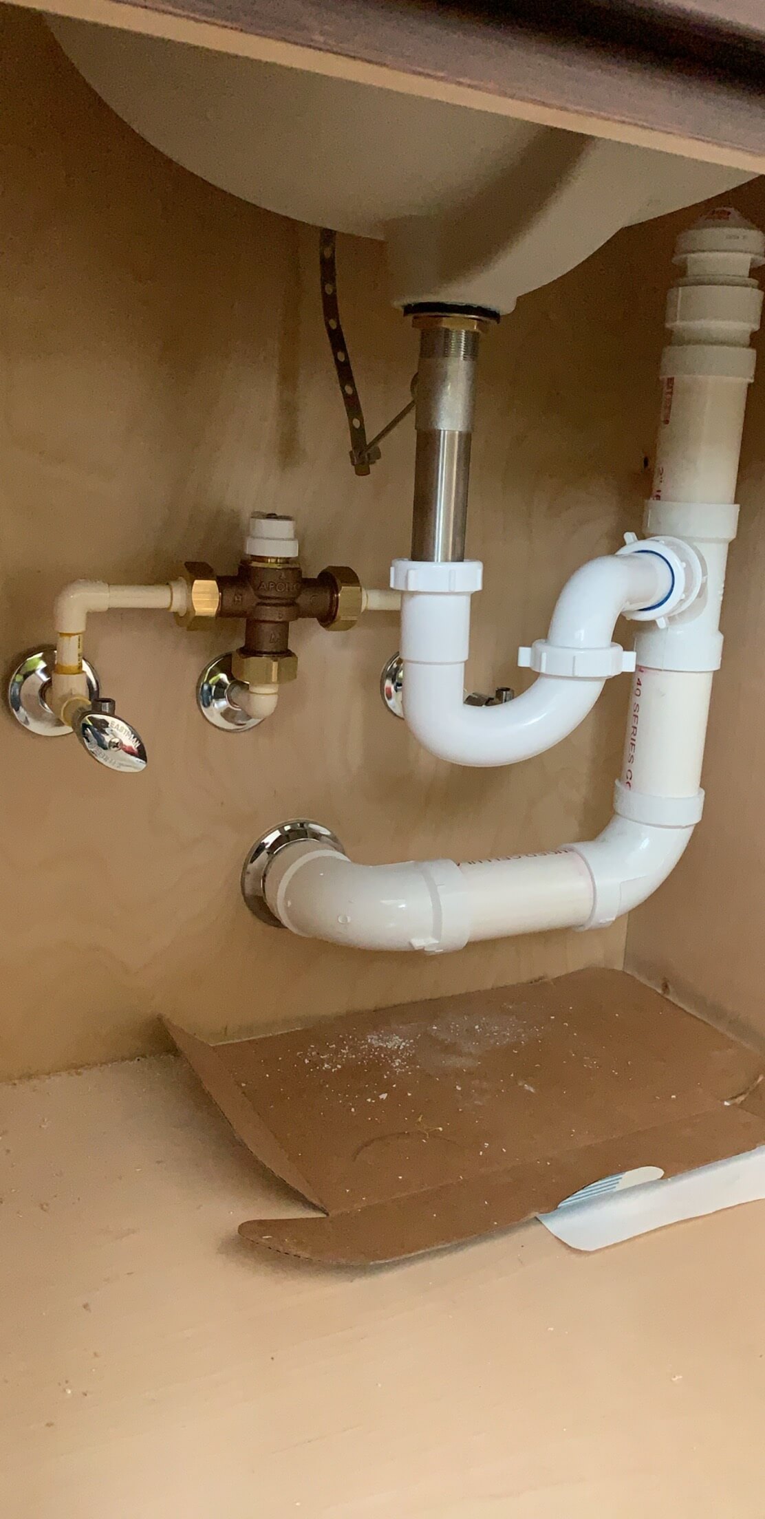 Plumbing pipes and valves under a sink inside a wooden cabinet with a piece of cardboard on the cabinet floor.