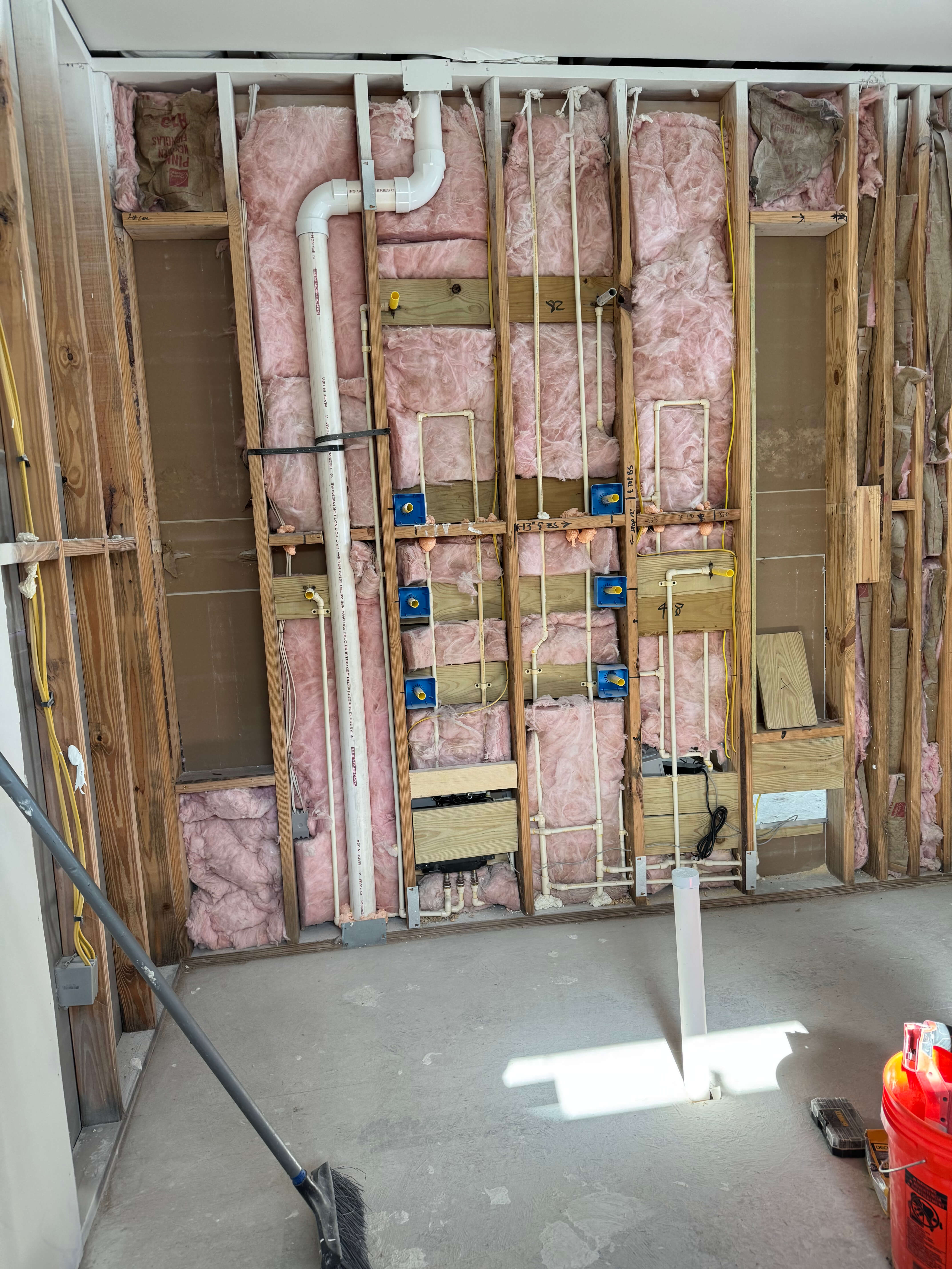 Interior wall under construction showing pink insulation, wooden studs, plumbing pipes, and electrical wiring.