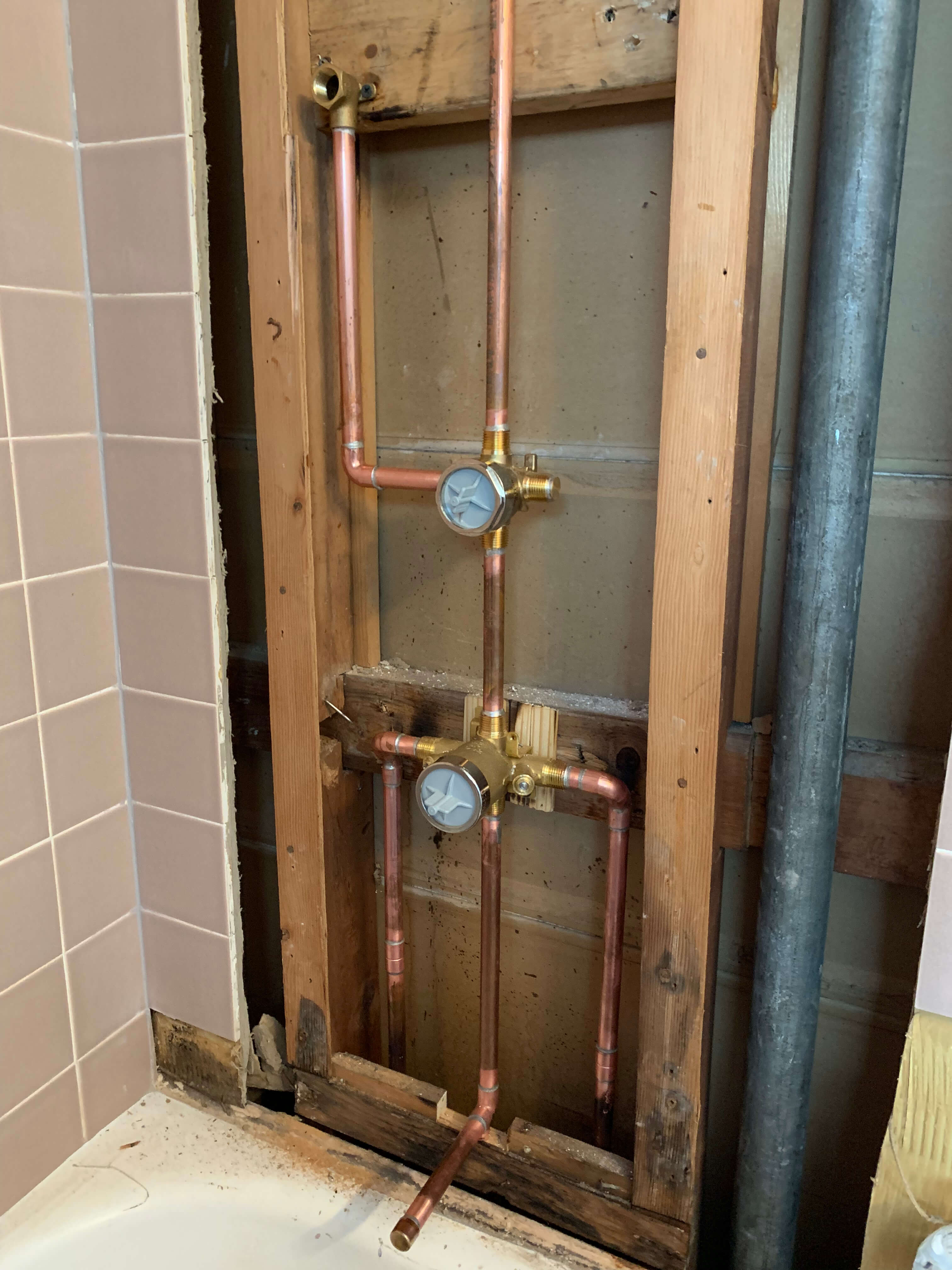 Exposed copper plumbing pipes and valves installed within a wooden wall frame next to a bathtub with beige tiles.